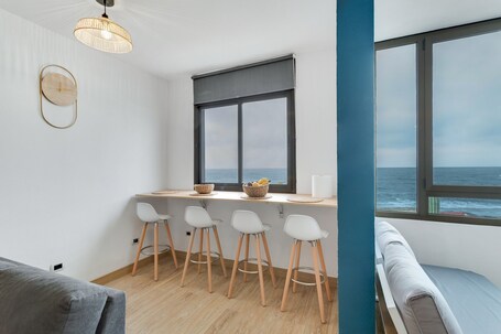 A modern room with a kitchen area, a dining set, a bench, and a large window with a view of the sea.. Vacation Apartment 'Casa Adrimar' with Sea View, Shared Pool and Wi-Fi