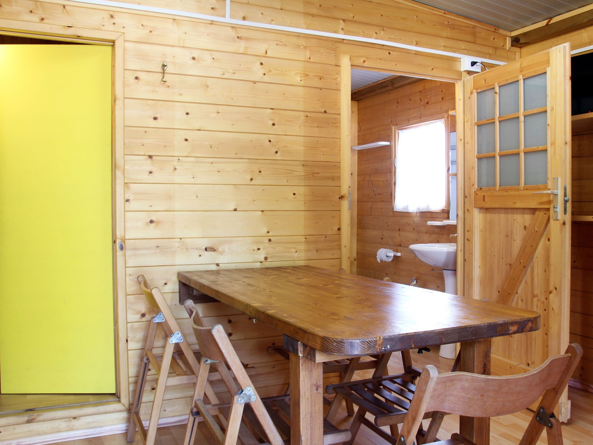 A wooden cabin interior with a dining table, chairs, a yellow door, and a window with frosted glass.