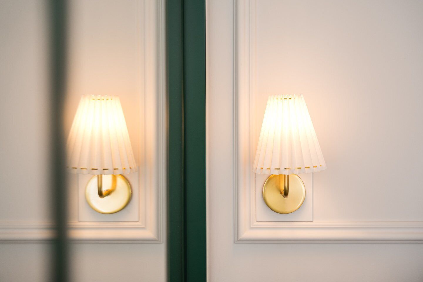 Zwei an der Wand befestigte Lampen mit klassischem Design, mit einem weißen Lampenschirm und einer goldenen Basis, an einer weißen Wand mit dekorativer Friese.
