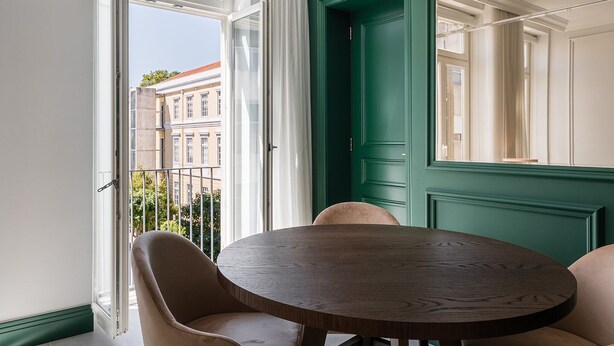A room with a round wooden table and two chairs, a green door, and a view of buildings through large windows. - GuestReady - Stylish stay with a balcony (Porto)