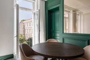 A room with a round wooden table and two chairs, a green door, and a view of buildings through large windows.