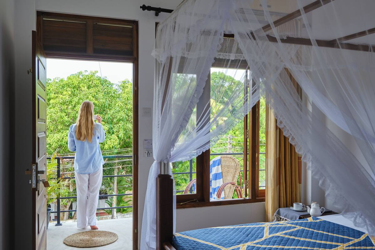 A bedroom with a bed, a window with a view of greenery, and a balcony with a chair.