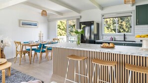 A modern kitchen with a large island, bar stools, and a dining area with blue chairs. - Racecourse Beach Coast Shack (Ulladulla)