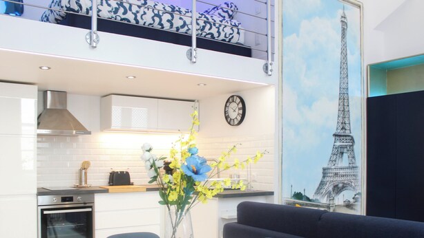 Une cuisine moderne avec des meubles blancs, un four encastré et un coin repas comprenant une table ronde et des chaises. Un vase rempli de fleurs est posé sur la table, et une horloge est accrochée au mur.