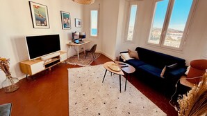 A modern living room with a flat-screen TV, a dark blue sofa, a small round table, and a large window offering a view of the city. - Charming Apartment at 10mn du Vieux Port (Marseille)