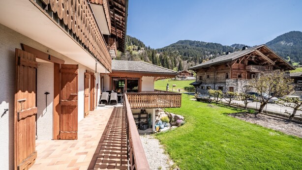 Un edificio de madera tradicional con balcón, un sendero pavimentado y vistas a un paisaje verde con montañas al fondo.