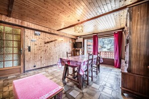 A wooden interior with a dining table, chairs, a television, and a view of the outside.