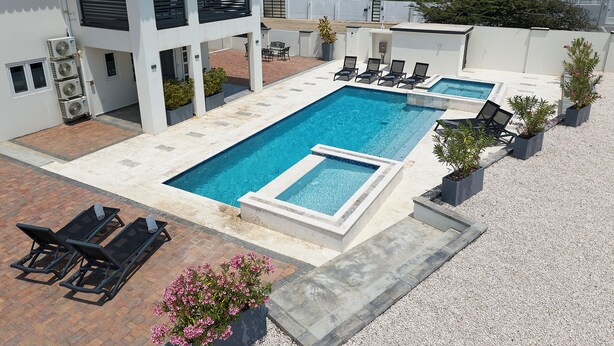 A rectangular swimming pool with a small adjacent hot tub, surrounded by lounge chairs and potted plants. - Kaikushi (Noord)