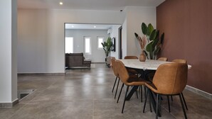 A spacious dining area with a marble table and brown chairs, a large potted plant, and a view into a living room with a sofa and a TV. - Kaikushi (Noord)