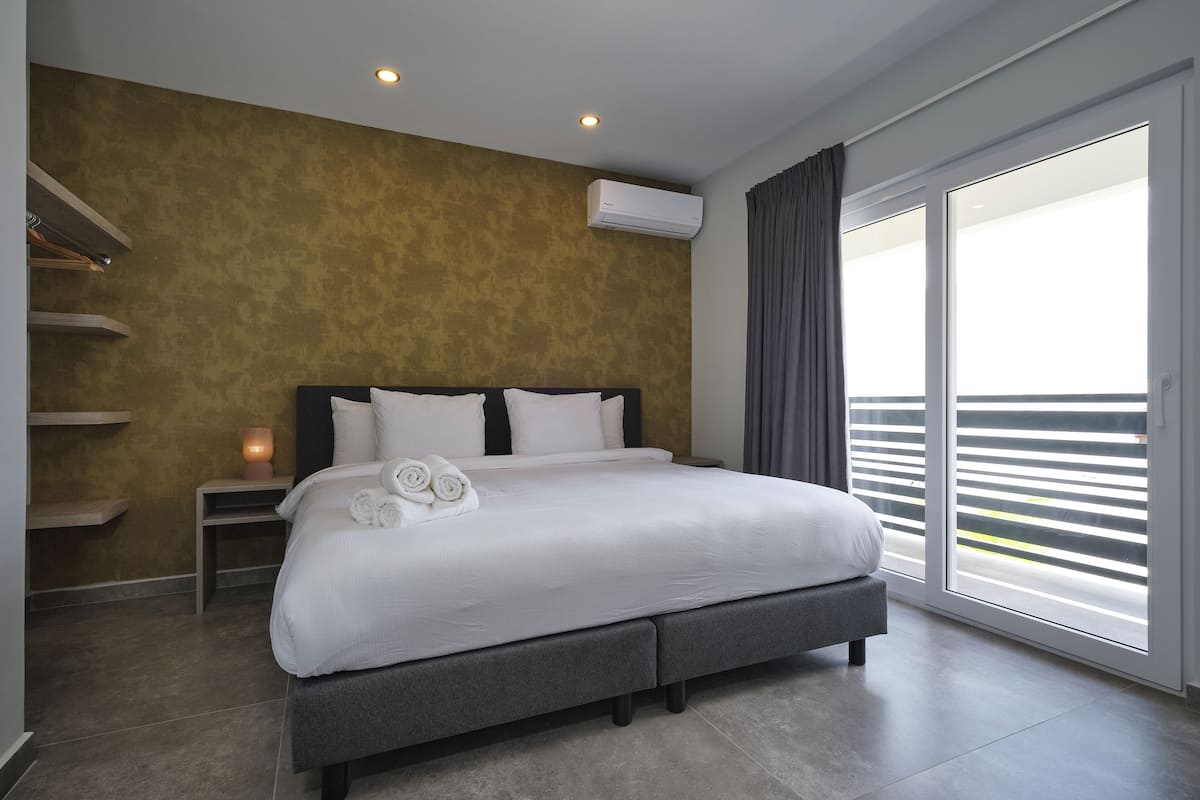 A modern bedroom with a large bed, a nightstand, a wall-mounted air conditioner, and a view through floor-to-ceiling windows.