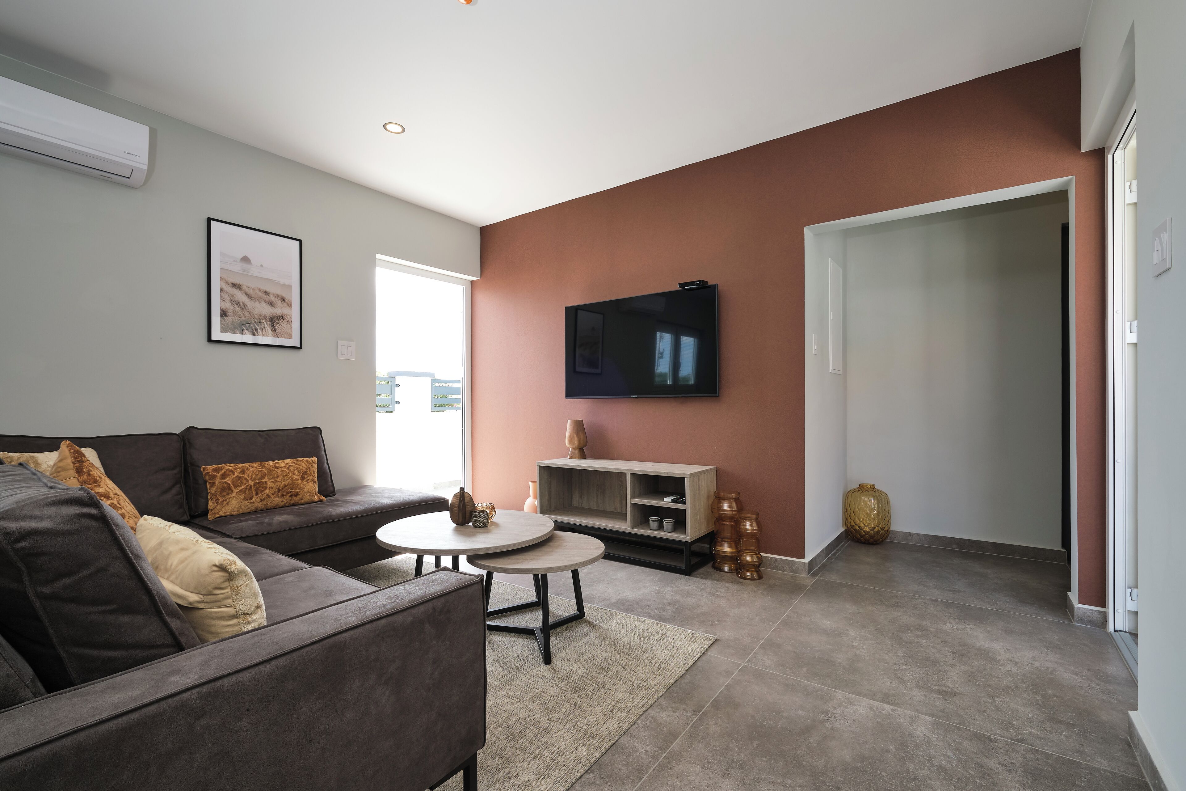 A modern living room with a grey sofa, two round coffee tables, a flat-screen TV mounted on the wall, and a large window providing natural light.