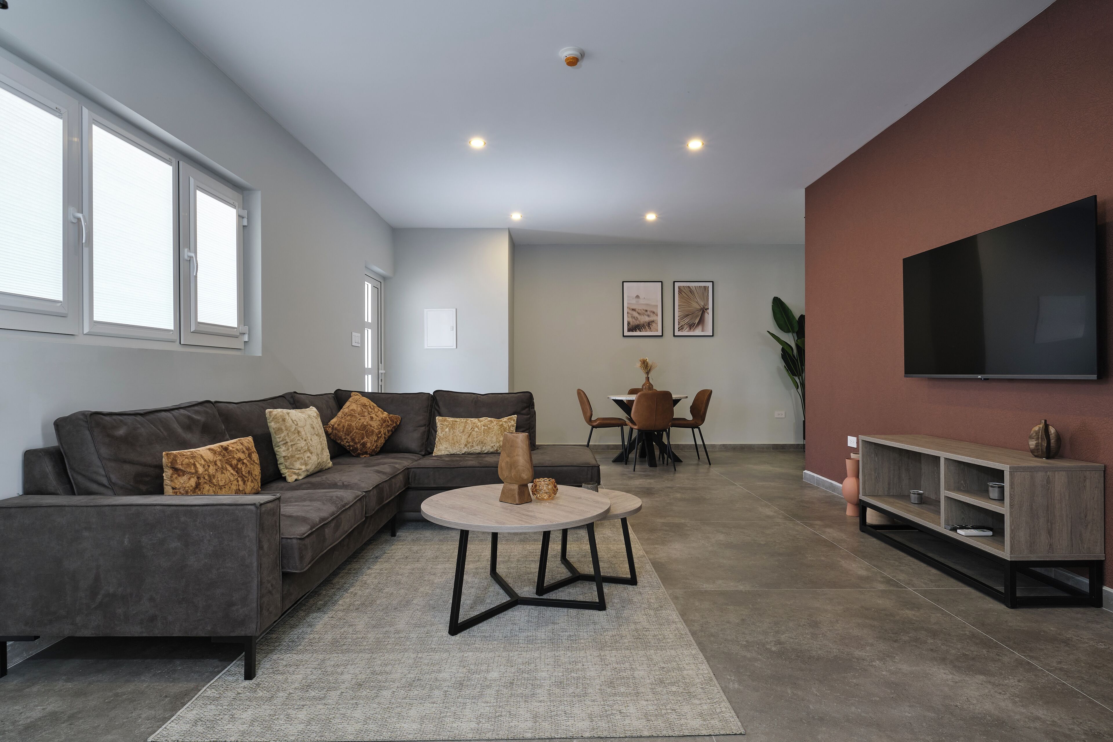 A modern living room with a grey sectional sofa, a round coffee table, a TV on the wall, and a dining area in the background.