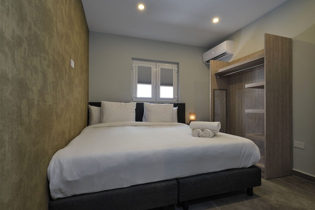 A neatly made bed in a room with a window, an air conditioning unit, and a wooden wardrobe.