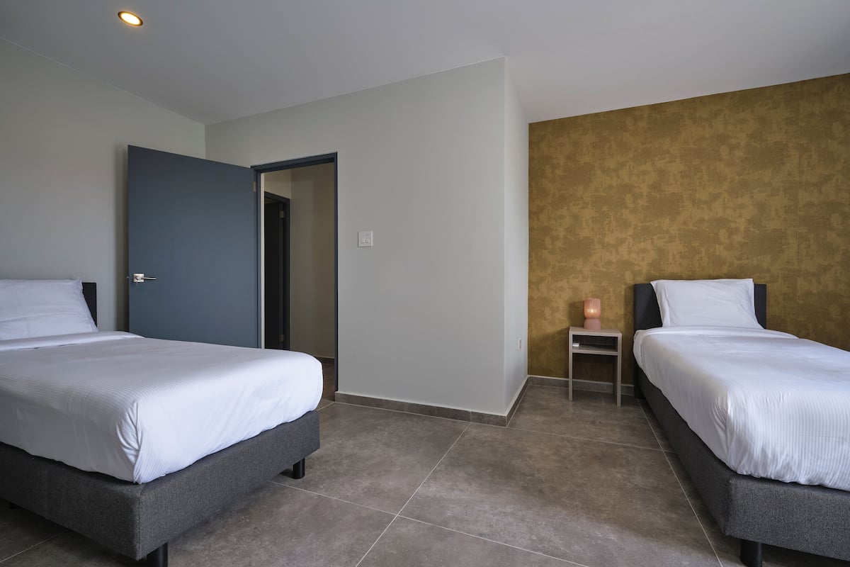 A hotel room with two single beds, a small bedside table, a nightstand, and a door leading to another room.