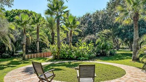 Un patio circular de ladrillo con dos sillas de madera y una mesa pequeña, rodeado de abundante follaje y palmeras.