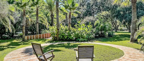 Un patio circular de ladrillo con dos sillas de madera y una mesa pequeña, rodeado de abundante follaje y palmeras.