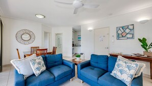 A modern living room with two blue sofas, a dining table, and a kitchen in the background. - Shoreline Apartments - Unit 15 (Bargara)