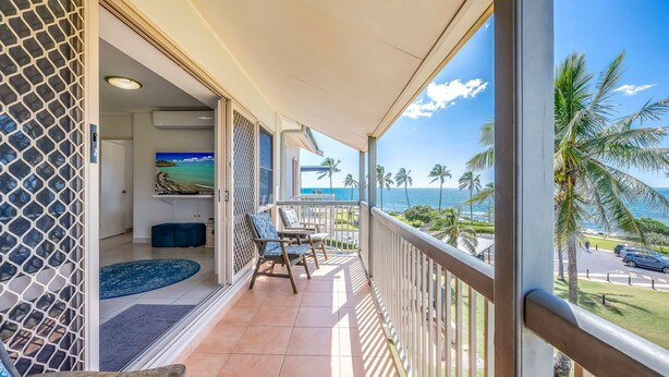 A balcony with a view of the ocean and palm trees, a TV on the wall, and a seating area with a blue rug. - Shoreline Apartments - Unit 15 (Bargara)