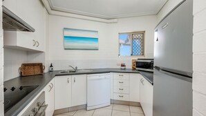 A modern kitchen with white cabinets, a dishwasher, a stove, a microwave, and a refrigerator. There is a window with a view of the outside. - Shoreline Apartments - Unit 15 (Bargara)