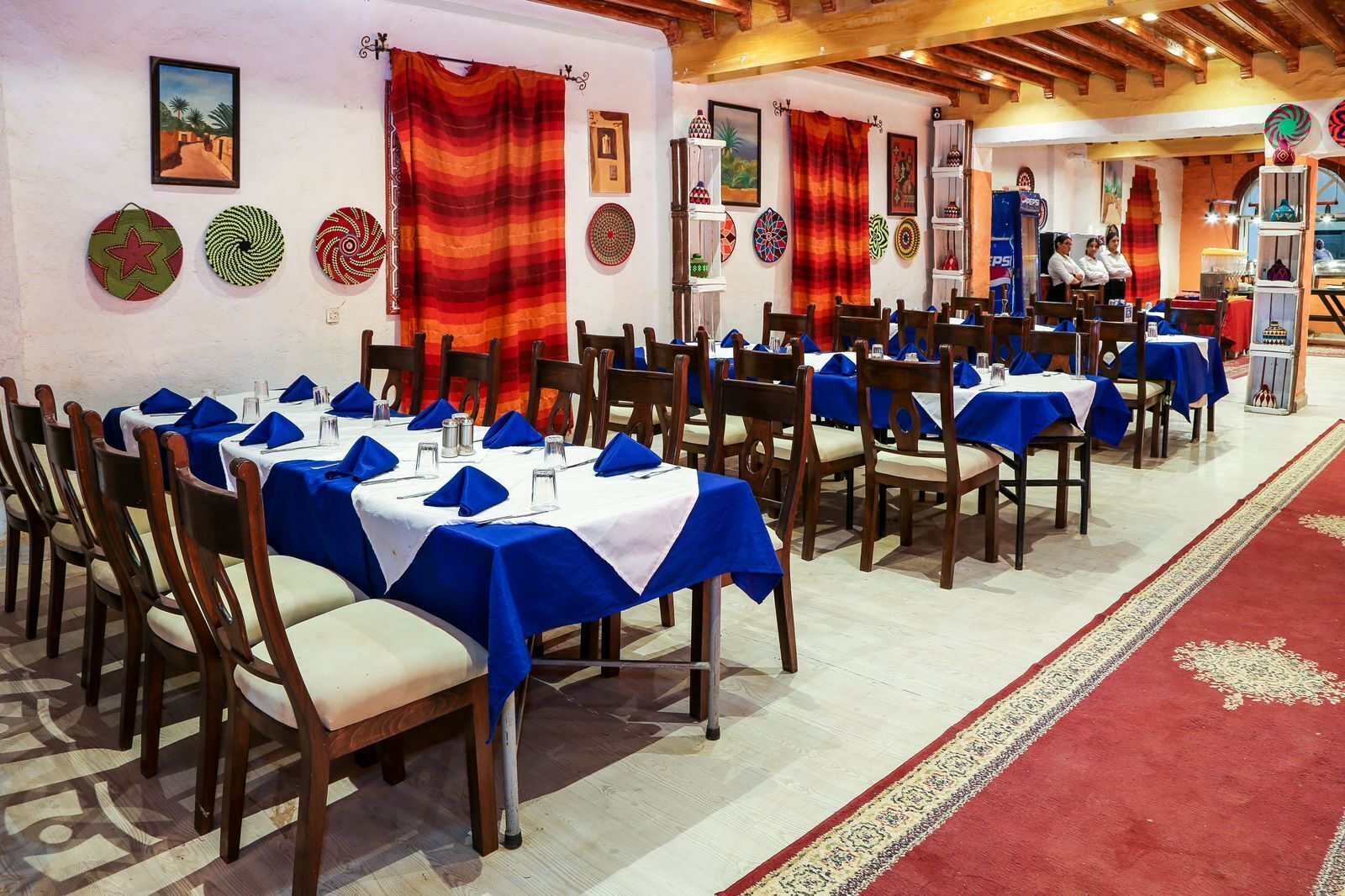 A restaurant with tables set with blue tablecloths and white napkins, wooden chairs, and a red carpet.