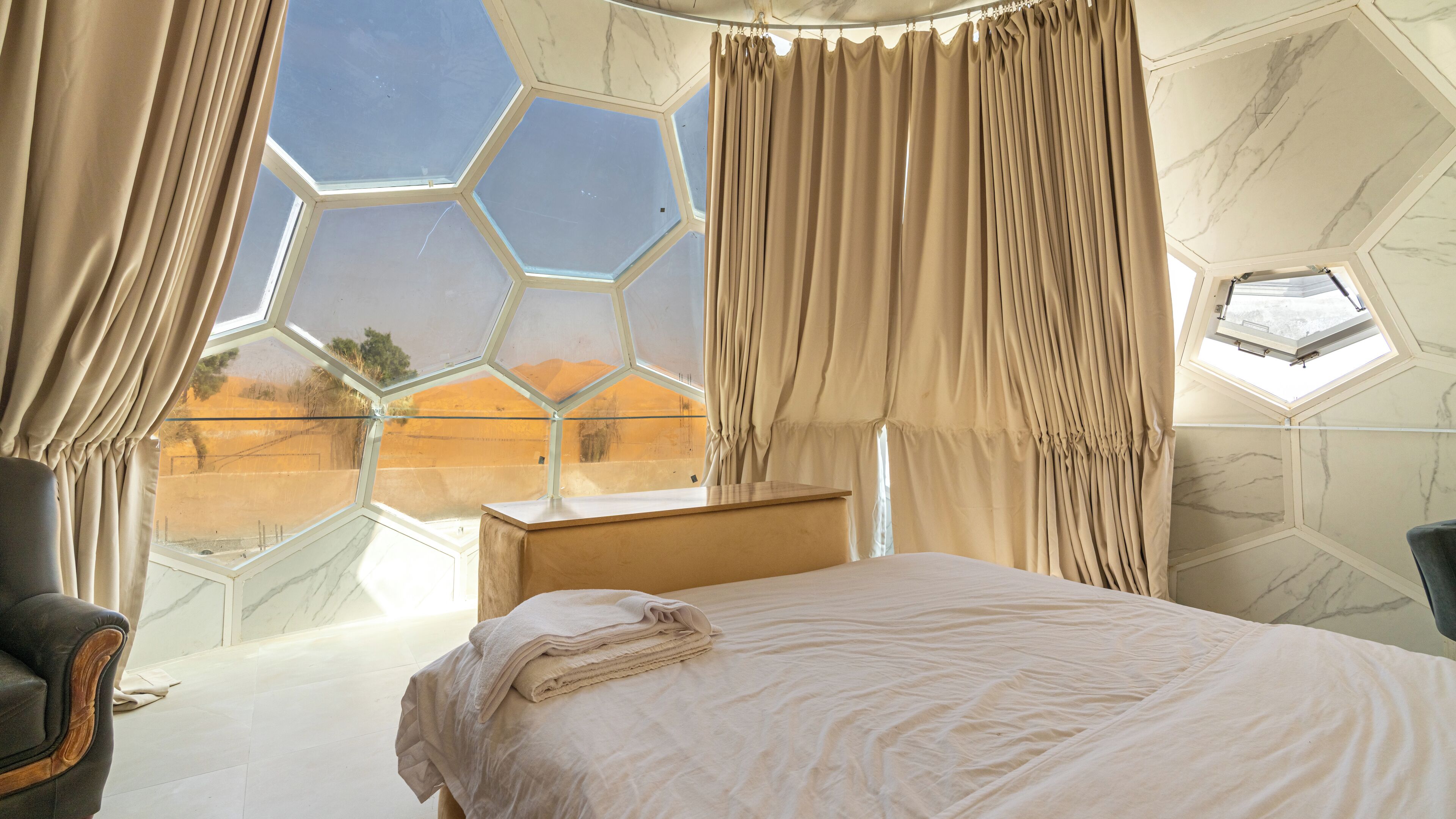 A bedroom with a large bed, a wooden nightstand, a chair, and a view through a hexagonal window.