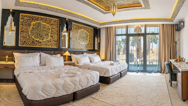 A hotel room with two beds, a desk, a TV, and a large window with a view of palm trees. - Kasbah Merzouga (Taouz)