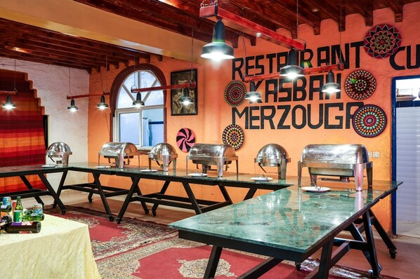 Un restaurante con una decoración rústica, que presenta una larga mesa comunal con calentadores metálicos para la comida, una alfombra roja y una pared decorada con platos.