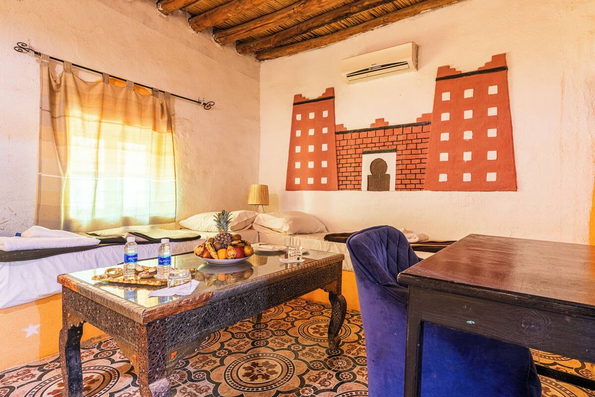 Une pièce comprenant un lit, une table avec un panier de fruits, une chaise et un mur dont le motif est en briques.
