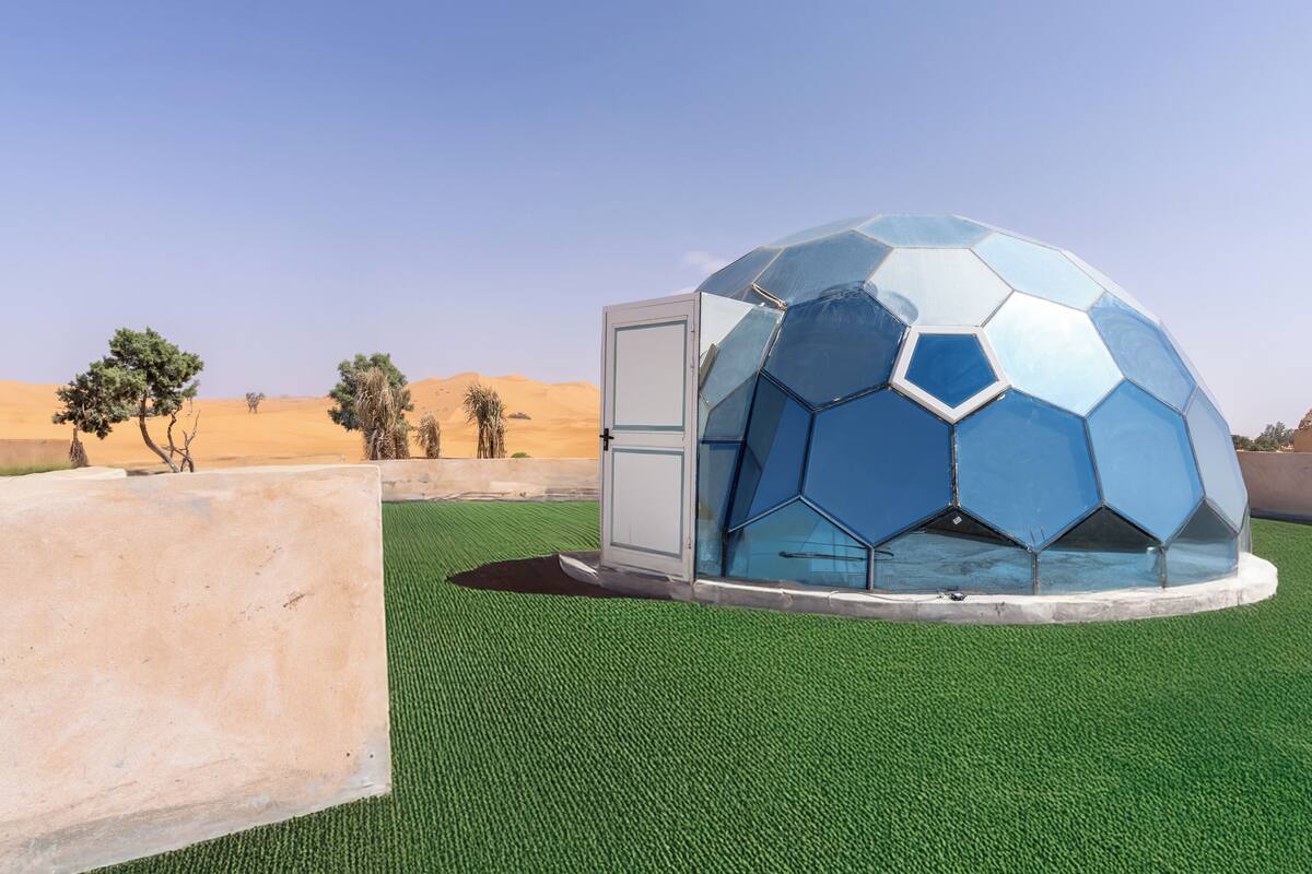 Une structure en forme de dôme, ornée d’un motif hexagonal, est installée sur un terrain recouvert d’une pelouse artificielle verte, avec un paysage désertique en arrière-plan.