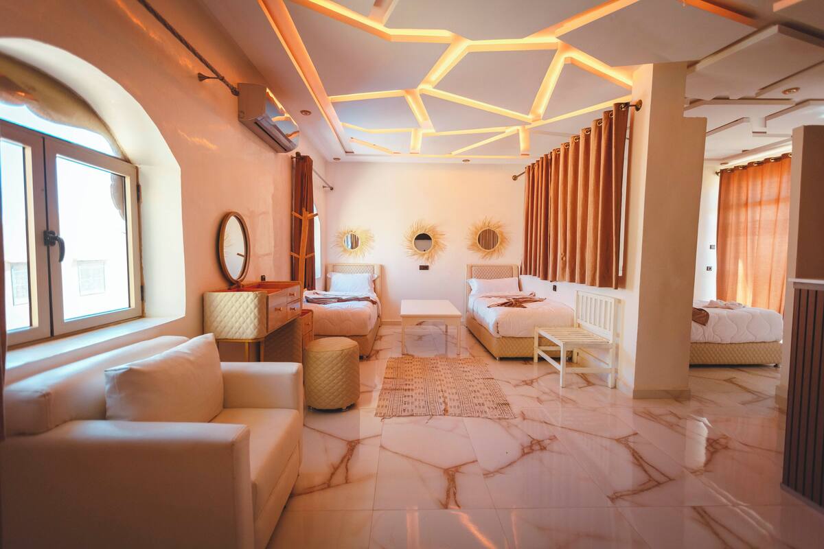 Une chambre d’hôtel moderne avec un sol en marbre, un canapé, un lit, une petite table et un plafond doté d’un éclairage aux motifs géométriques.