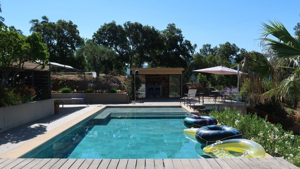Ein rechteckiger Pool mit Holzterrasse, umgeben von üppiger Vegetation und einer überdachten Sitzgelegenheit.