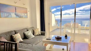 Ein Wohnzimmer mit grauem Sofa, einem weißen Couchtisch und Meerblick durch große Schiebeglasfenster.