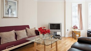 Un salon avec une table basse en verre, un canapé rouge, un téléviseur et une fenêtre avec des rideaux.