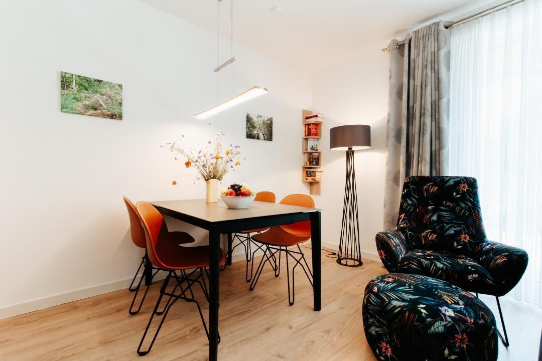 Ett modernt vardagsrum med en matplats, med bland annat ett trägolv, ett svart matbord med orange stolar, en svart fåtölj med ett blommönster och en golvlampa.