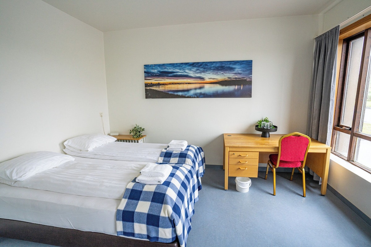 A hotel room with a bed, a desk, a chair, a window, and a painting on the wall.