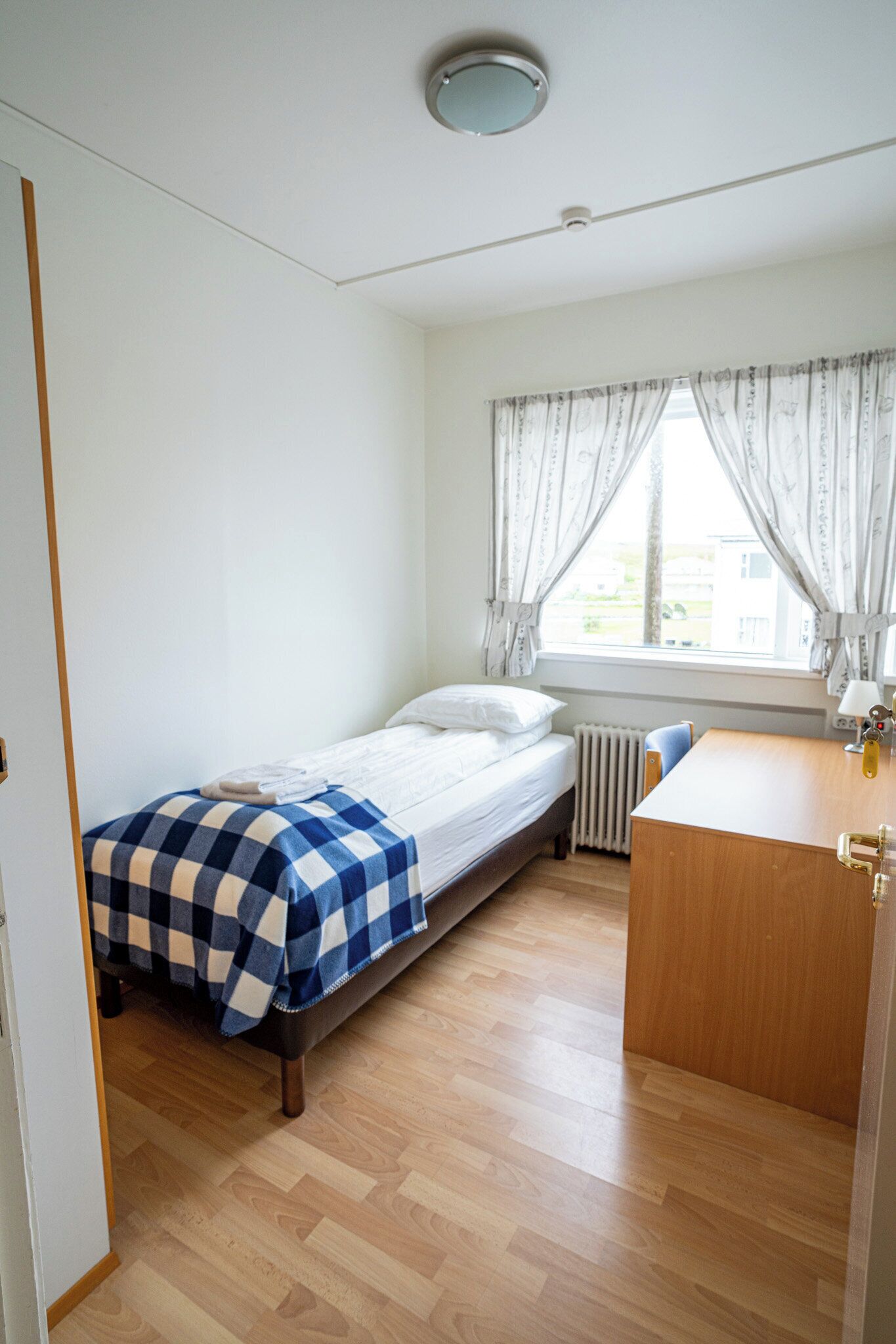 Une petite pièce bien éclairée, avec un lit simple, un bureau en bois et une fenêtre avec des rideaux.