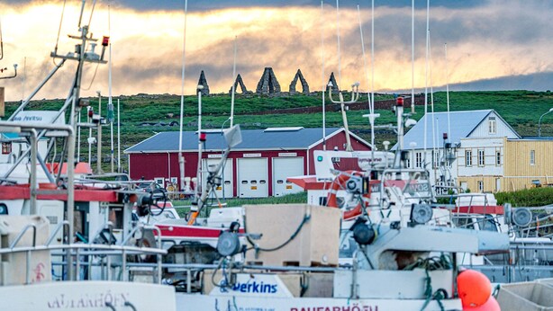 Een haven met boten, een rood gebouw en een heuvel met stenen constructies op de achtergrond.