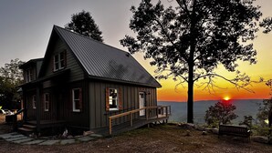 A rustic cabin with a steep gabled roof, a front porch, and a view of the sunset over a body of water.