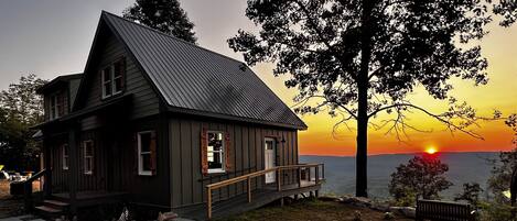 A rustic cabin with a steep gabled roof, a front porch, and a view of the sunset over a body of water.