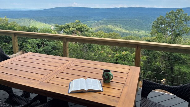 A wooden table with an open book and a mug, overlooking a mountainous landscape.
