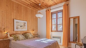 A bedroom with a wooden ceiling, a bed with a white bedspread, two bedside lamps, a framed picture on the wall, a window with curtains, and a radiator. - Le Prunier by Interhome (Oraison)