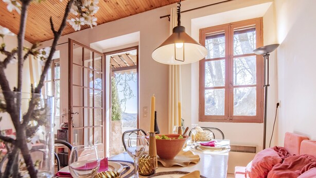 A dining area with a table set for two, a vase with flowers, and a view of the outdoors through a window. - Le Prunier by Interhome (Oraison)