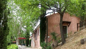 A gravel path leading to a brick building with a red door and windows. There are outdoor tables and chairs under a red umbrella, and a staircase leading up to the building. - La Chênaie by Interhome (Oraison)