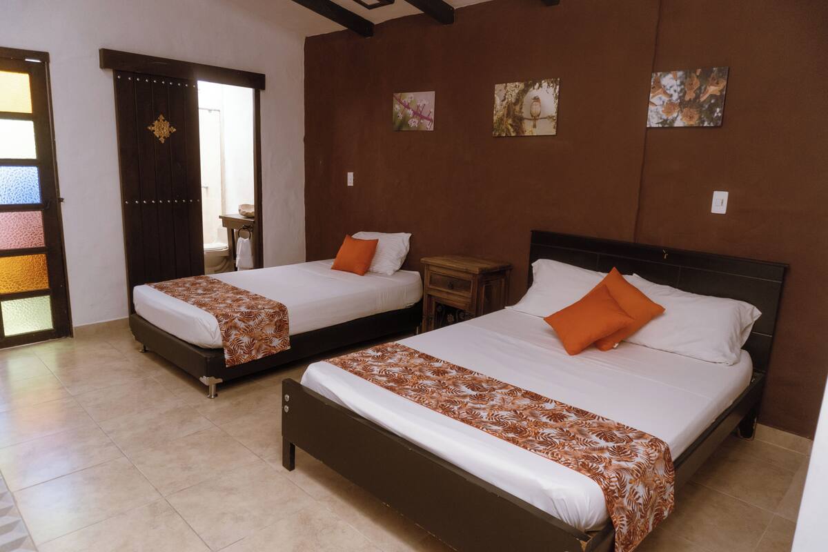 A hotel room with two beds, each with a brown headboard and white bedding. There are wooden nightstands and a small table between the beds. The walls are adorned with framed artwork, and the room has a tiled floor.