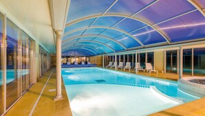 Een binnenzwembad met een gebogen plafond en glazen wanden, omringd door loungestoelen.