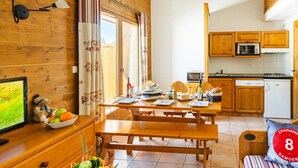 Une pièce chaleureuse avec une table en bois, des bancs, un lit, un téléviseur et un coin cuisine doté de meubles et d’appareils électroménagers en bois.