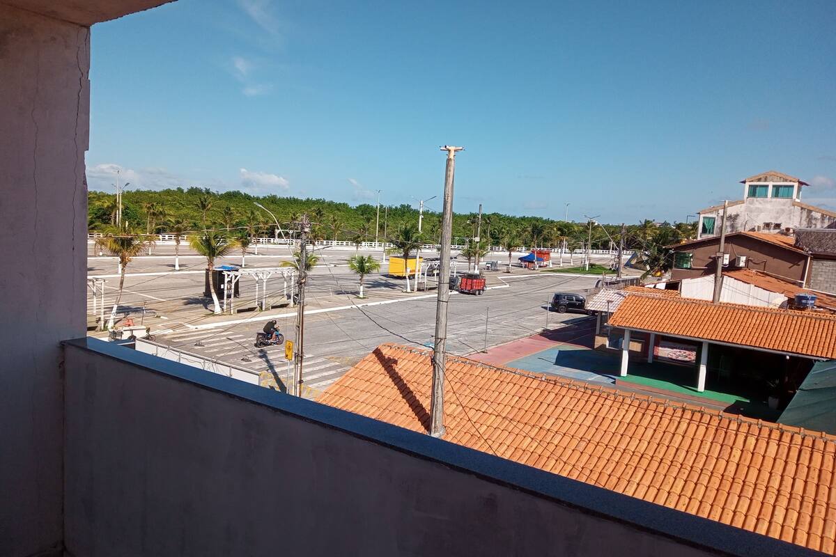 A view from a balcony overlooking a street with a red-roofed building, a parking lot, and a green area with trees.