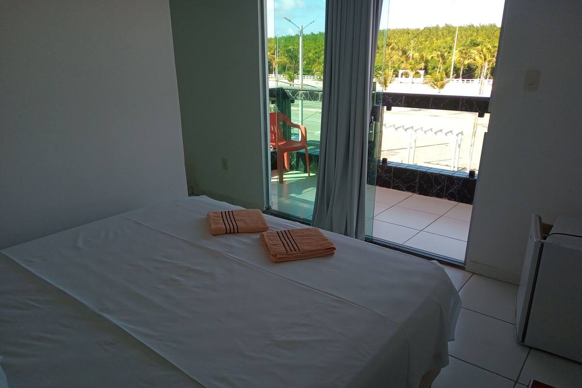 A hotel room with a bed, two towels, a window, and a view of a balcony with chairs and greenery outside.