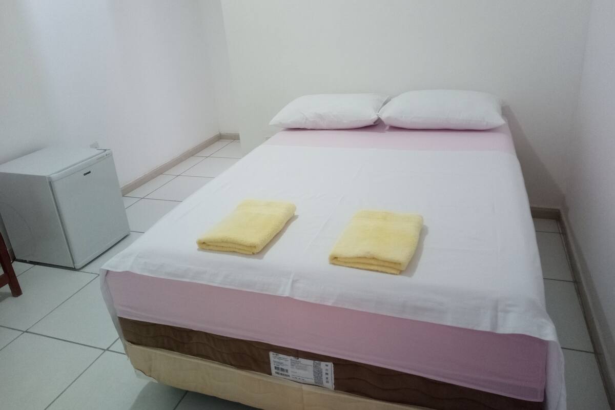 A small, neatly made bed with two folded towels, a white pillow, and a pink bedspread in a minimalistic room with white walls and a tiled floor.
