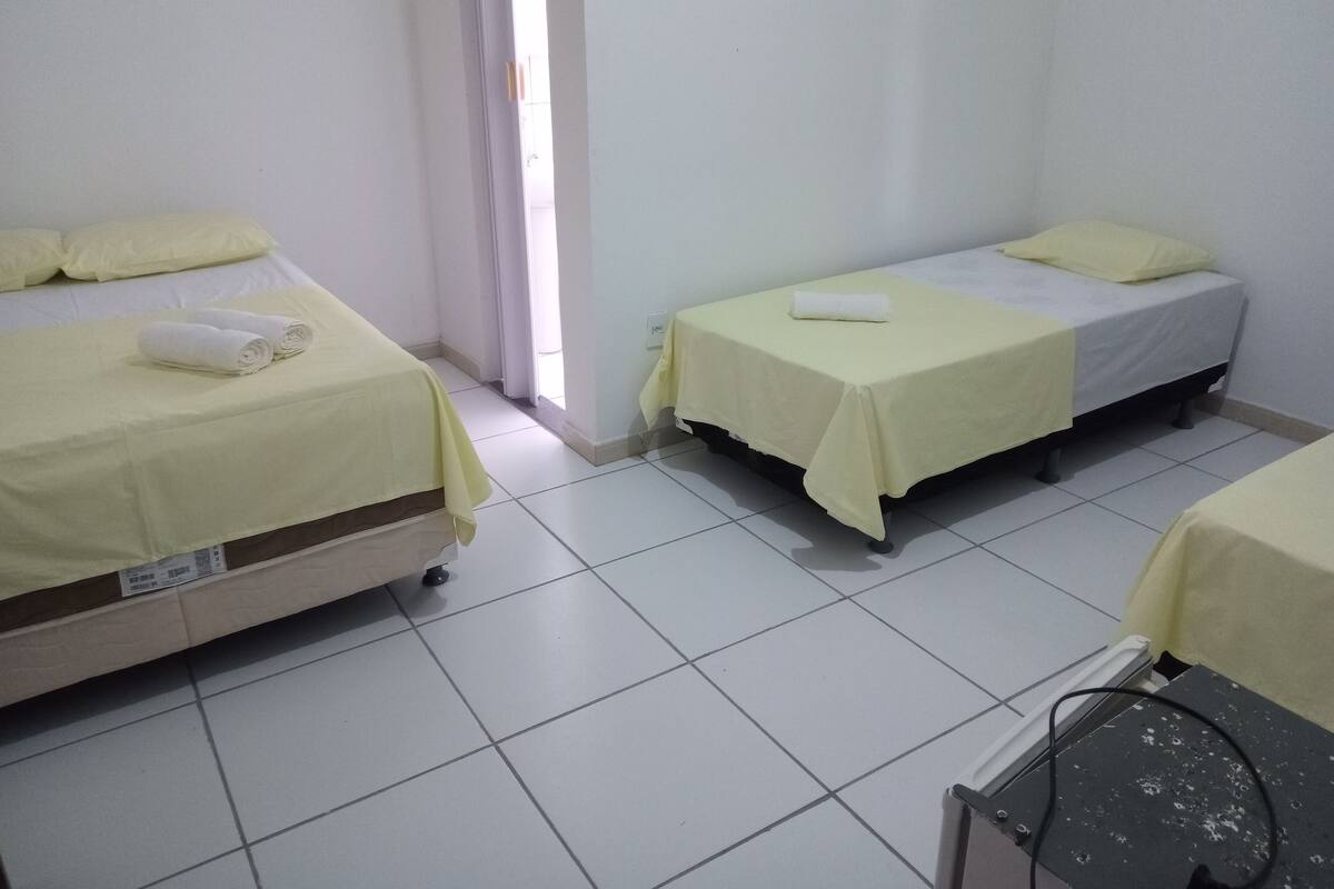 A hotel room with two beds, each with a yellow bedspread and pillows. There is a window with curtains, a bedside table with a phone, and a tiled floor.